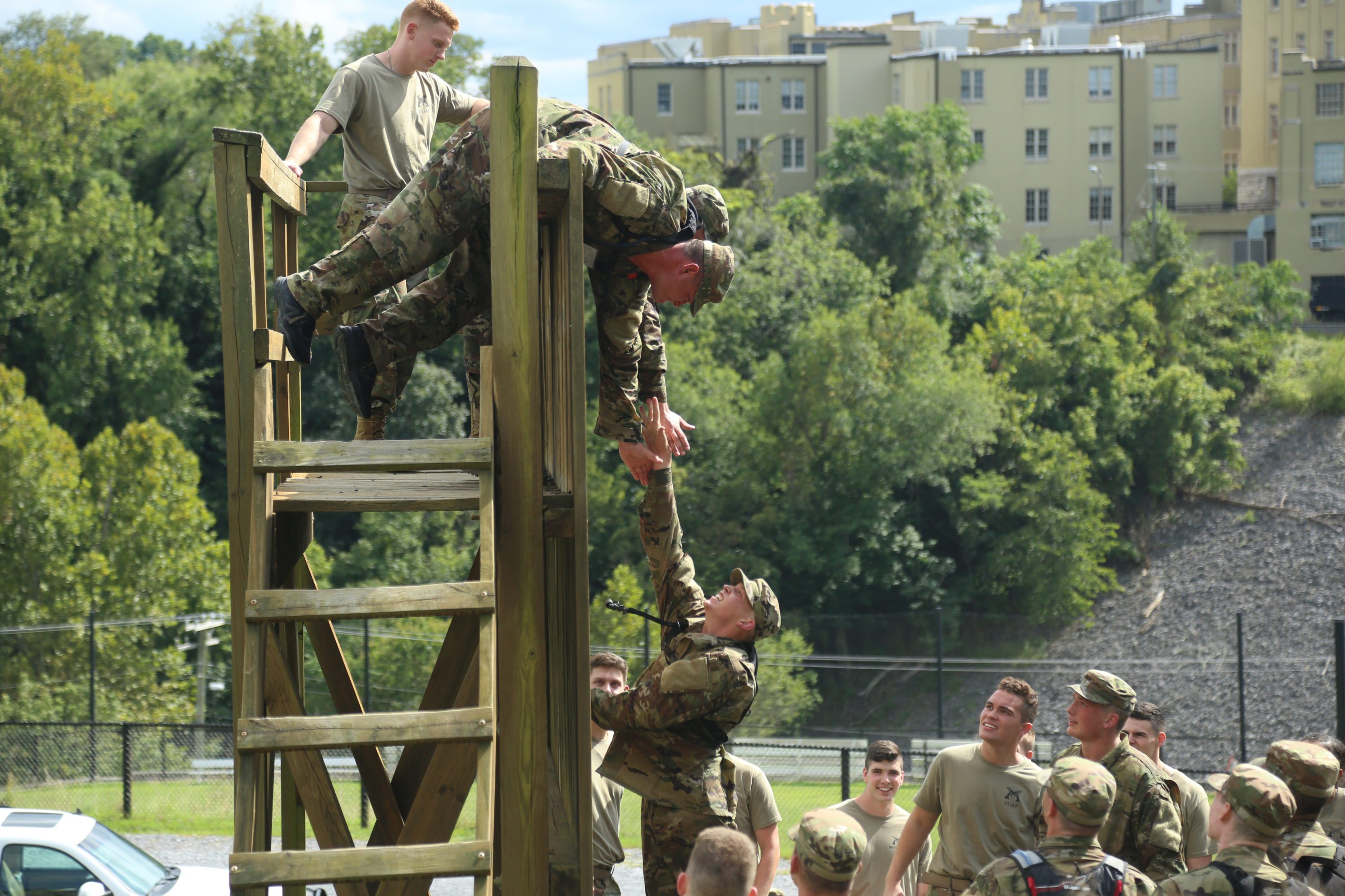 two cadets reach down to a fellow cadet to help him climb wooden structure