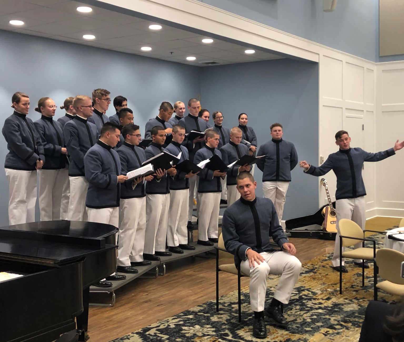 Glee club standing on risers, with one member seated in a chair, singing and smiling.