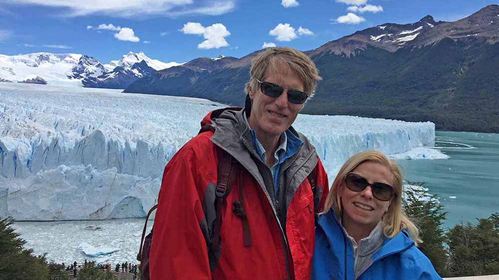 Man and woman stand in front of glacier, smiling.