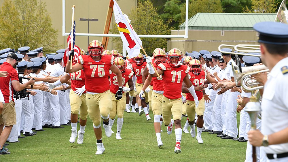 Football team running onto field, surrounded by cadets in uniform.