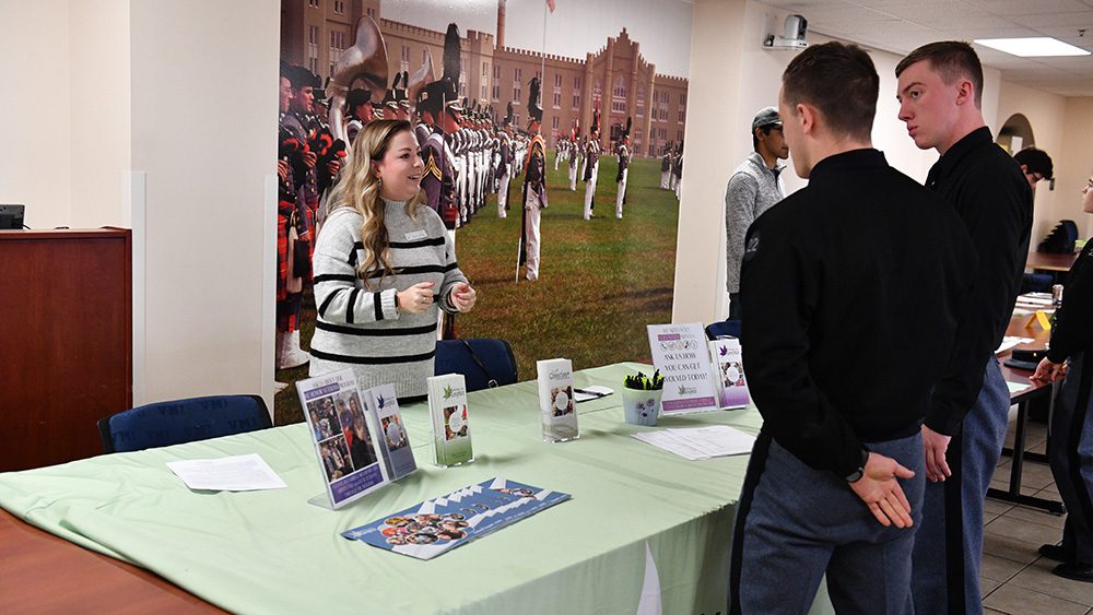 woman stands behind table with pamphlets on it, talking to two cadets