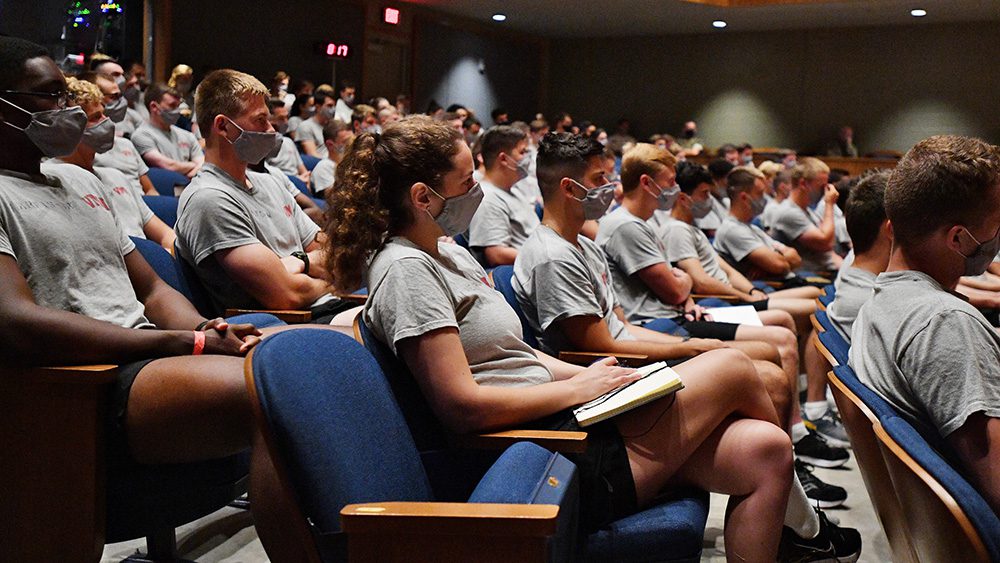 cadets in gym dyke and masks seated in auditorium taking notes