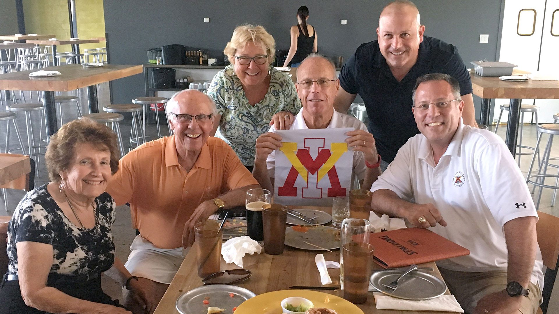 Group of men and women seated around a table, holding up a VMI flag, smiling.