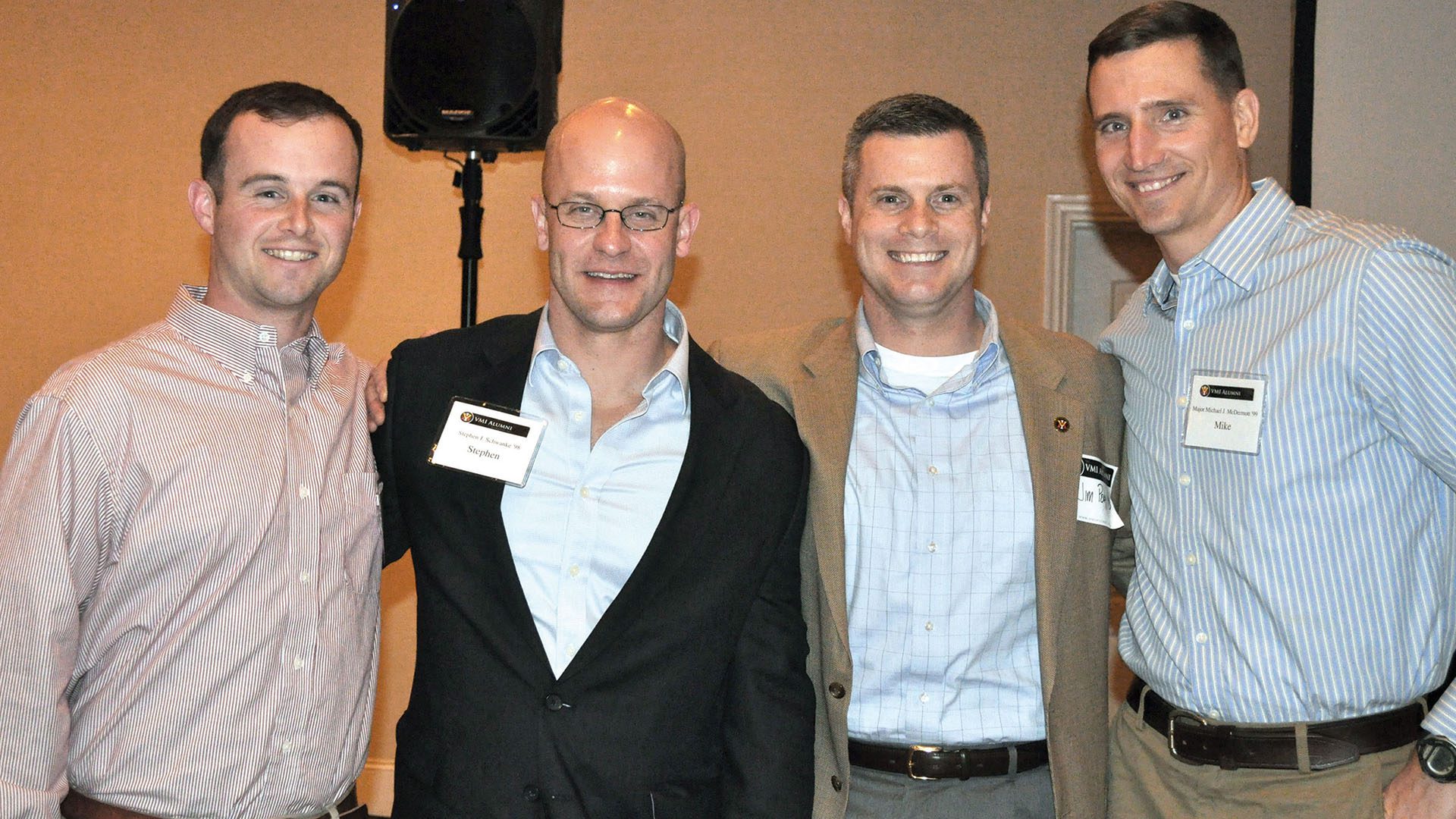 Four men wearing nametags pose with arms around each other, smiling.