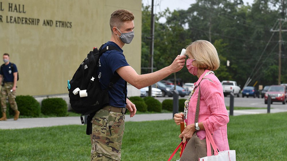 male cadet wearing face mask using forehead thermometer to take woman's temperature