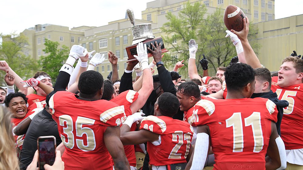 football team cheering, surrounding Maj. Gen. Wins '85 holding Silver Shako trophy over his head