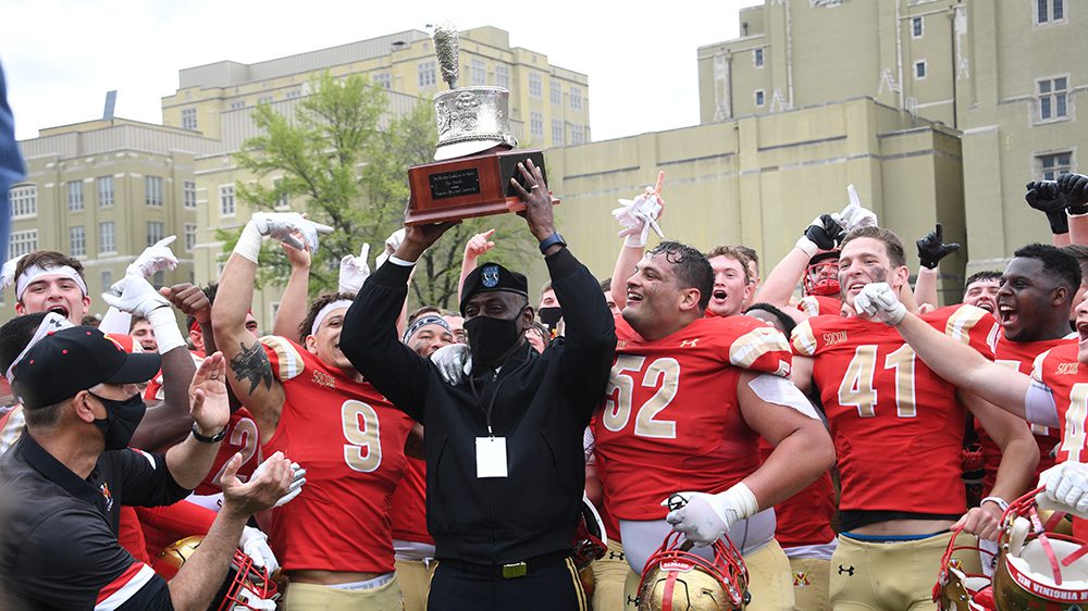 football team cheering, surrounding Maj. Gen. Wins '85 holding Silver Shako trophy over his head