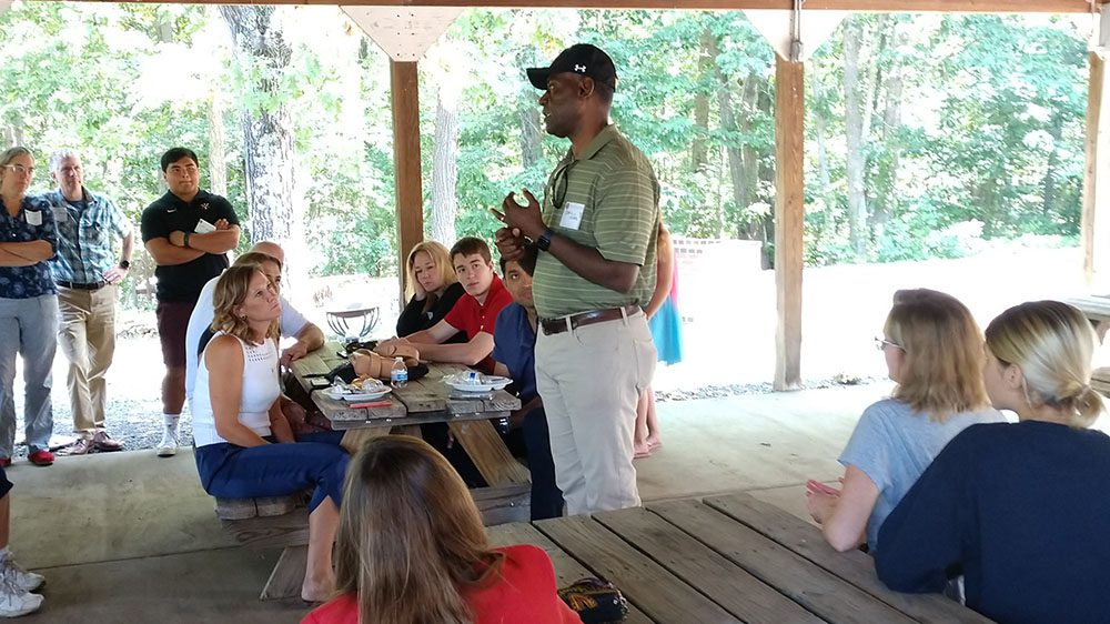 Man standing, speaking to a group of people seated at picnic tables.