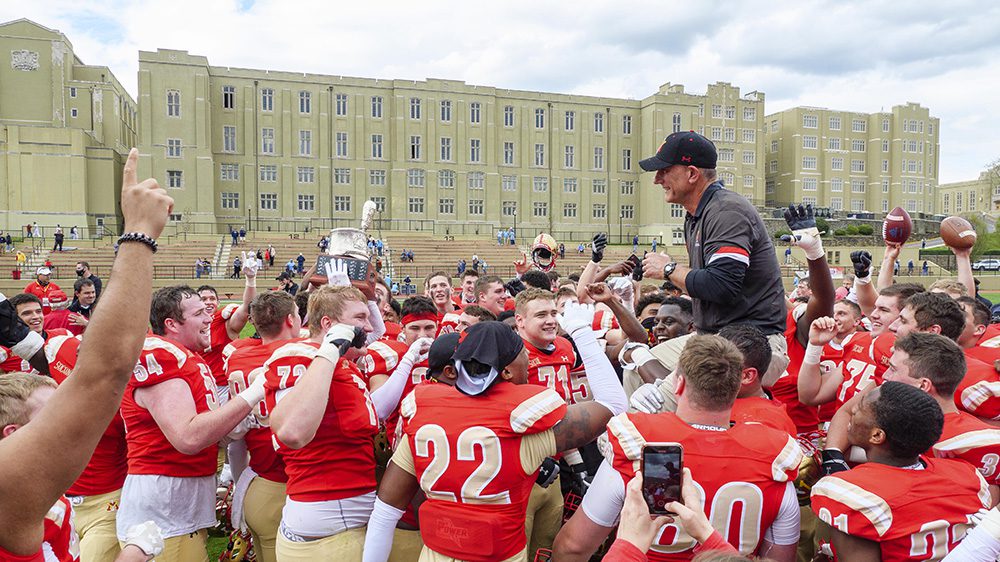 Keydet football team hoisting Coach Scott Wachenheim on shoulders and cheering, while one player holds Silver Shako trophy.