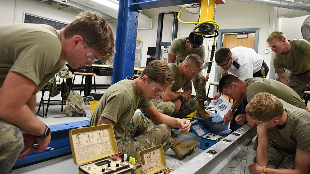 Col. Matt Swenty assists cadets in his civil engineering lab as they sit or kneel on the floor and set up gauges before stress testing an I-beam.