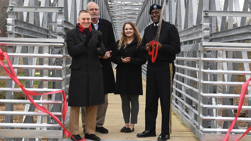 Four people pose on Chessie Trail bridge smiling, Maj. Gen. Wins holding giant red scissors