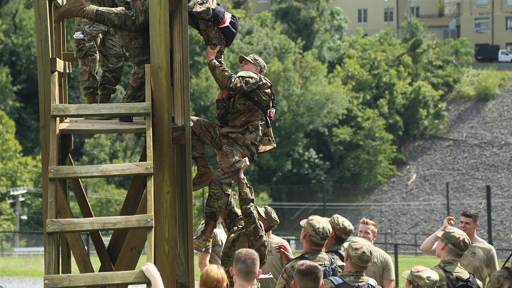 one cadet reaching down to help another cadet over a wooden structure, while a group of cadets boosts the second cadet up
