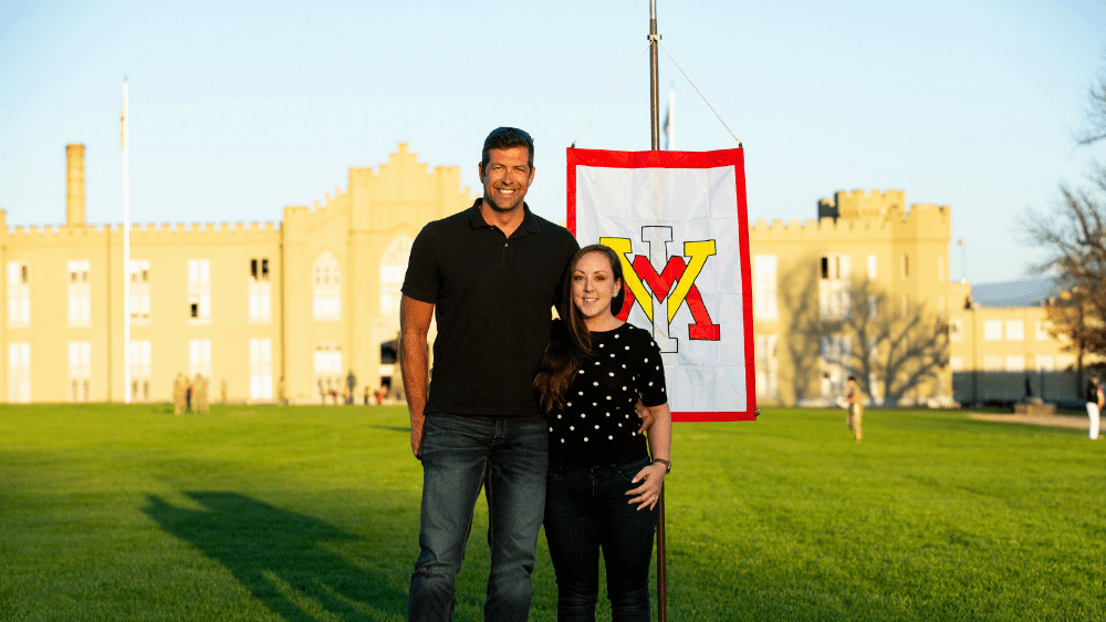 man and woman posing in front of barracks and VMI flag, smiling