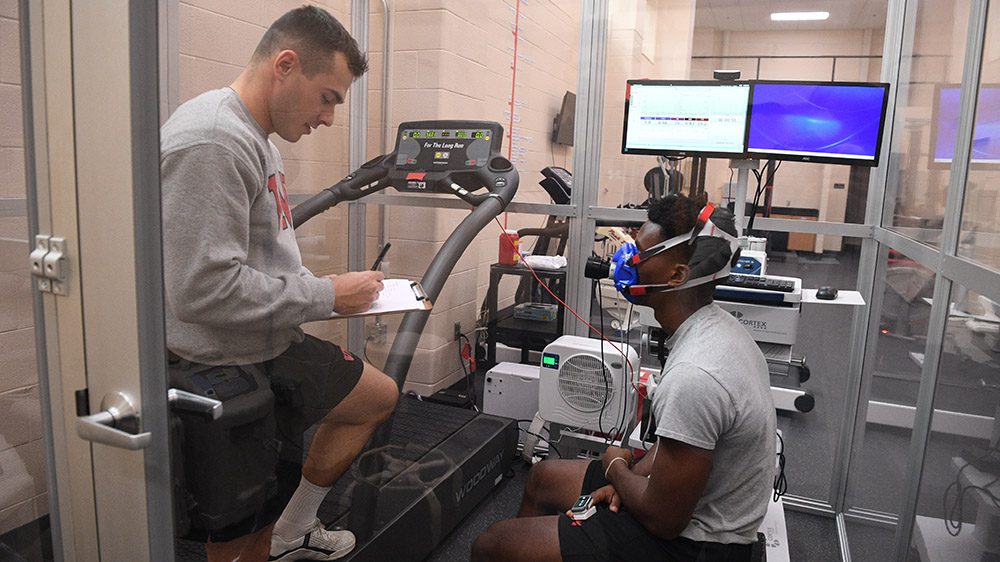 Two cadets sit in altitude chamber, one taking notes, while the other sits with mask over nose and mouth.