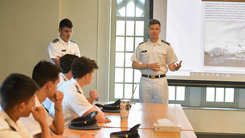 Cadet stands in front of projector screen, addressing a group of cadets seated at a table.