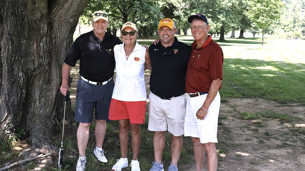 Three men and a woman pose outside with arms around each other, smiling.