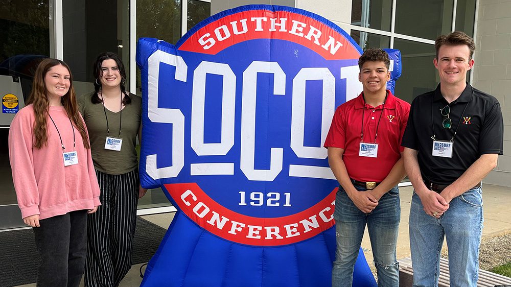 Cadets Caragh Osborne ’24, Gabriella Handford ’24, Anthony Fernandez-Grimes ’25, and William Davidson ’24 posing in front of SOCON logo sign.