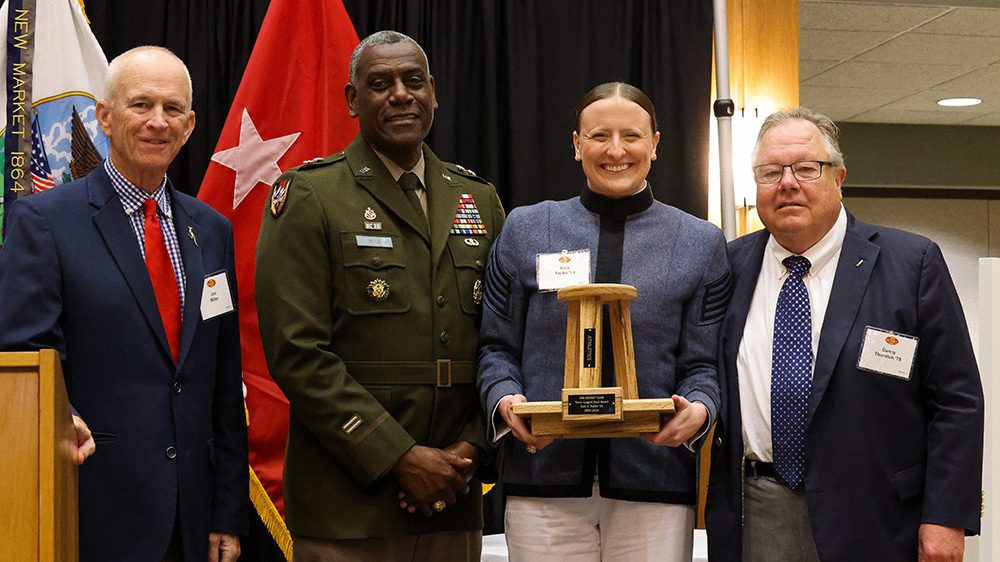 Jim Miller, Maj. Gen. Cedric T. Wins, Cadet Kate Taylor, and Danny Thornton smiling, as Taylor holds Three-Legged Stool trophy.