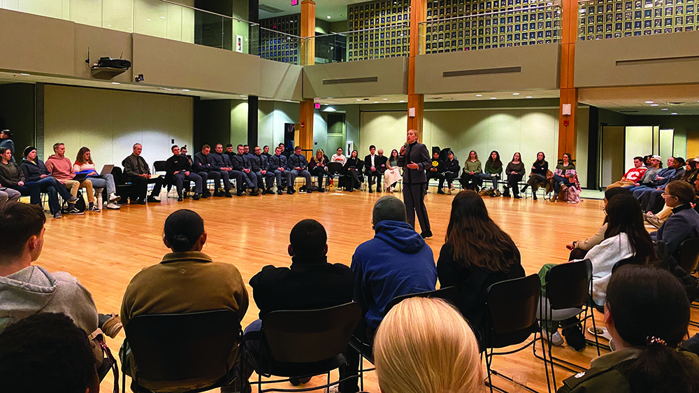 female cadet speaking into a microphone with audience seated in a circle around her