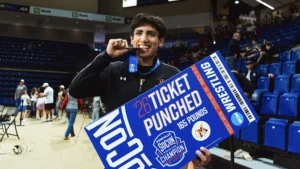 wrestler holding poster that says "'26 Ticket Pubched, 165 pounds, Asheville SoCon Wrestling Champion" and biting his medal.