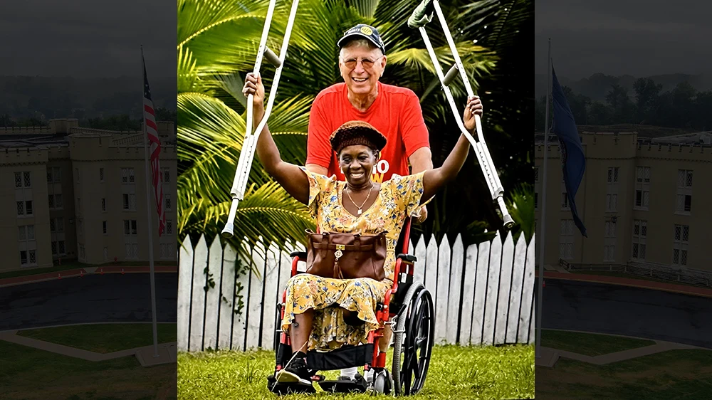 A woman sits in a wheelchair and smiles, holding up crutches, while Woody Sadler ’66 pushes wheelchair