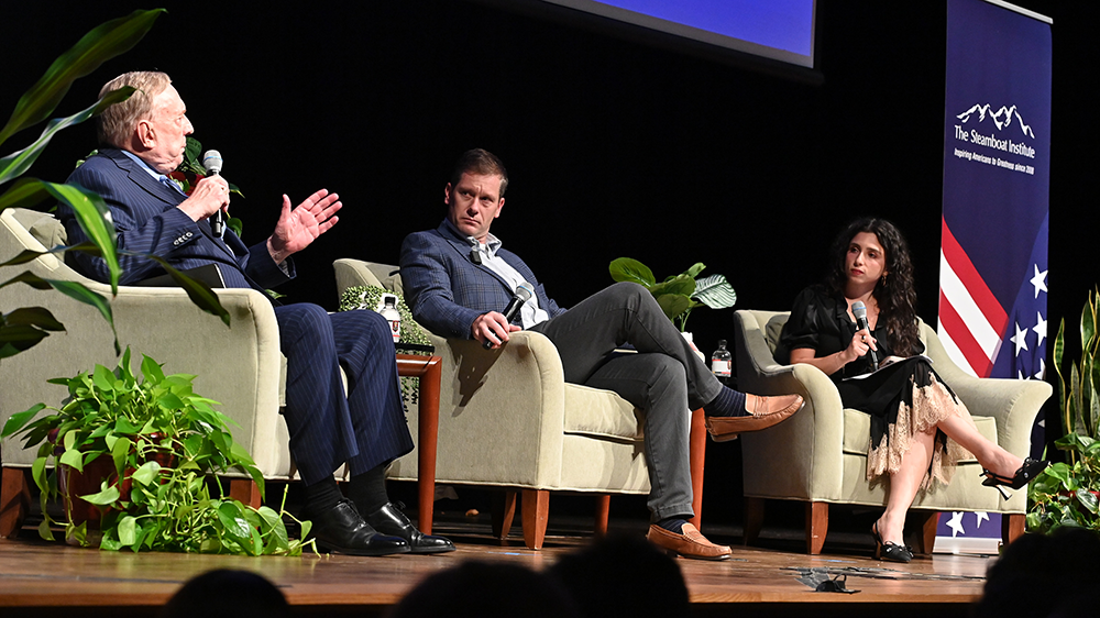 two men and one woman sit in chairs on stage in the CLE. One man is speaking into a microphone while the others listen.